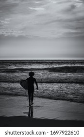 Lone surfer walks into the ocean to catch a wave. View on an Atlantic coast and dramatic cloudy sky at sunset. Concept of outdoor activities. Lisbon, Portugal.High Contrast. B&W color.

