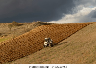A lone tractor plows through the fields, storm clouds looming above.  A solitary tractor tills a vast field, its lines of progress marked under the ominous gaze of gathering storm clouds.