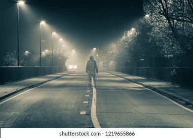A lone transparent ghostly hoodied figure standing in the middle of a foggy road at night. With a vintage duo tone edit.