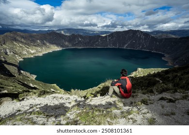 A lone traveler gazes out over the breathtaking Quilotoa Crater Lake in Ecuador. The vibrant turquoise waters of the lake contrast against the rugged, mountainous landscape, creating a serene. - Powered by Shutterstock