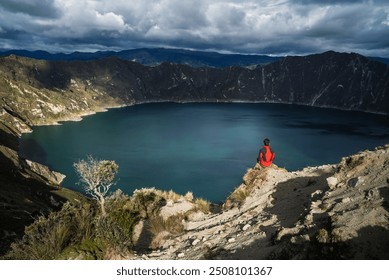 A lone traveler gazes out over the breathtaking Quilotoa Crater Lake in Ecuador. The vibrant turquoise waters of the lake contrast against the rugged, mountainous landscape, creating a serene. - Powered by Shutterstock