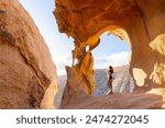 A lone traveler stands gazing at the natural arch of Arco de las Penitas, set against the rugged backdrop of Fuerteventuras landscape in the golden afternoon sunlight.