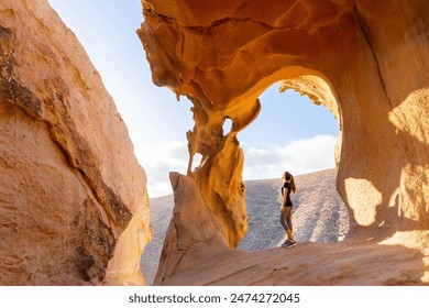 A lone traveler stands gazing at the natural arch of Arco de las Penitas, set against the rugged backdrop of Fuerteventuras landscape in the golden afternoon sunlight. - Powered by Shutterstock