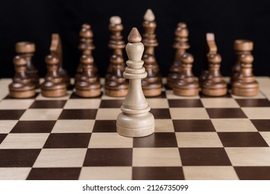 A lone white chess king stands on the chessboard opposite the opponent's chess pieces. The concept of the courage of a leader challenging an opponent. One warrior in the field. One against the enemy.