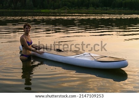 A lone woman rides a Sup Board on a lake surrounded by grass and dense green forest at sunset. Lake Lebyazhye, Kazan. Summer landscape.