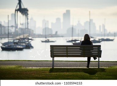 Lone woman in silhouette sitting on a bench by the waters edge with boats and city skyline in the background.