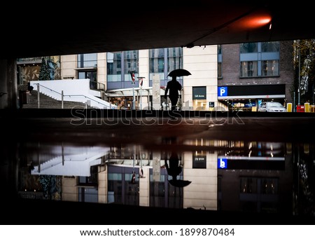Lone worker man walking through underpass with umbrella up commuting to work in modern city centre flood water puddle reflected. Rainy day reflection dark silhouette bad severe weather big puddle