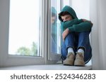 Loneliness concept. Sad teenage boy on windowsill indoors