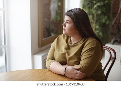 Loneliness concept. Young brunette plus size woman with black hair sitting at cafe table, feeling lonely, spending time alone, waiting for her lunch, looking through window with sad pensive expression