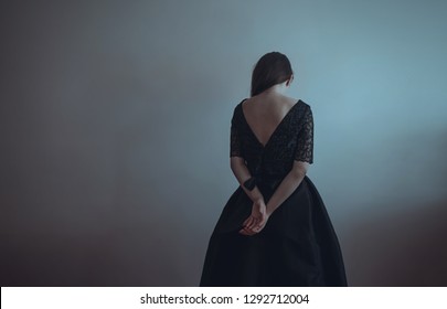 Loneliness. Lonely girl stands near the gray wall. Solitude, sadness, depression, disappointment. Woman in vintage black dress. Girl is afraid and hugs herself. Dramatic, painful picture. Pain.