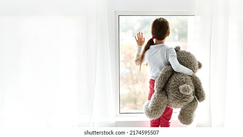 Loneliness. Lonely Orphan Girl In An Orphanage Looking Outside While Holding Teddy Bear The Window. Child Feelings Concept