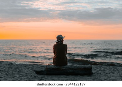 Lonely adult woman wearing a coat and hat sitting alone on a log by the sea at sunset looking away, back view. - Powered by Shutterstock