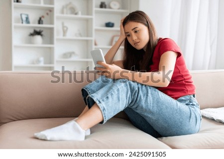 Lonely asian woman with smartphone sitting on sofa at home, upset young korean lady looking at mobile phone screen, waiting for message, having depression, relaxing in living room interior