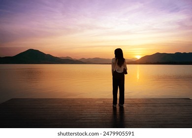 Lonely Asian woman standing on the wooden bridge watching sunset sky in the evening. Back of silhouette single sad teenager girl standing alone on the waterside. Lam Taphen Reservoir, Suphanburi. - Powered by Shutterstock