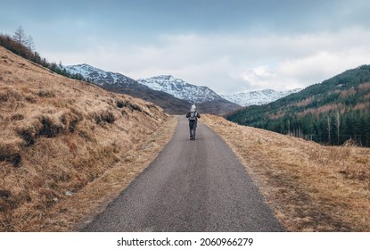 Lonely Backpacker Man Walking With Backpack By Scottish Highlands Mountain Road With Snowy Peaks At Early Moody Morning. Active People In Beautiful Nature Concept Image. Scotland, United Kingdom.