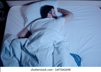 A Lonely Bearded Dark-haired Man Sleeps Soundly Lying On His Stomach At Night On The Bed, Hands Clasped. The General Plan From Above, White Bedding, A Bedroom, Self-isolation. One