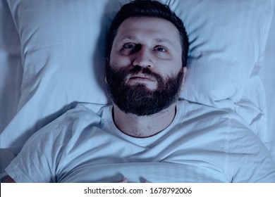 A lonely bearded man in bed at night cannot sleep, is unhappy and very tired. Sleep problems, stress, insomnia, bedroom, pajamas, white bedding.Close up - Shutterstock ID 1678792006