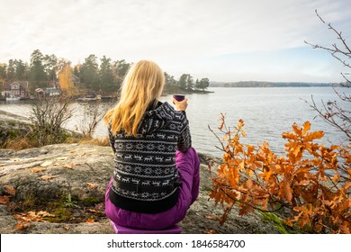 A lonely blonde girl sits on top of a rocky cliff and looks at the panorama of the sea bay. Woman traveler tourist hiker with a mug of hot tea on the shore. Archipelago with forest islands in autumn.