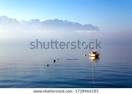 Lonely boat on Lake Leman or Lake of Geneva with morning mist over the water surface. At the background are the snow-covered Alps in Montreux, Switzerland