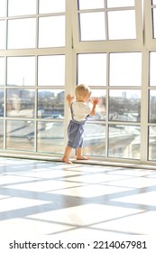 A Lonely Child And An Abandoned Little Boy Stands At The Window And Looks Out The Window In The Rays Of The Sun Waiting For His Mother