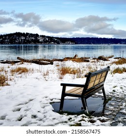 A Lonely, Empty Park Bench Beside A Snow Covered Walkway On A Bright Winter Day With A View Over The Ocean Of A Distant Island And Clouds In The Sky 