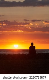 A Lonely Figure Of A Woman Sitting On The Seashore At Sunset. The Sun Is Sinking To The Horizon, Beautifully Coloring The Sky And Water In Golden Pink Tones. Copy Space.           