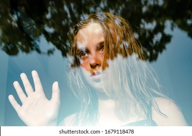 Lonely Girl Due To Self Distancing At Home During Corona Virus. Quarantine From Covid-19 World Pandemic. Anxiety Child Looking Worried Through Window Glass Waving  