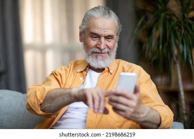 Lonely Handsome Bearded Senior Man Sitting On Couch At Home, Using Brand New Cell Phone, Touching Gadget Screen And Smiling, Using Dating Mobile Application, Copy Space