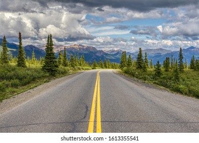 Lonely Highway Heads Through The Tundra Towards The Mountains Under Storm Clouds In Alaska.