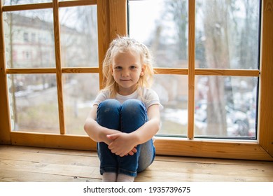 Lonely Little Blonde Girl Sitting On A Wooden Windowsill Near Big Window In New House. 