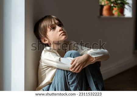 Lonely little boy sadly looking up hugging knees sits near window at home alone. Upset unhappy child waiting for parents, thinking about family problems, bad relationship. Kid psychological trauma