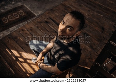 A lonely man sits on a chair in an old room. Looking into the camera with his blue eyes, wrinkles are visible on the man's forehead. The shadow from the window falls on the floor.