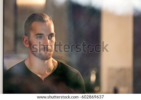 Lonely man wearing a black long sleeved shirt, standing in his home looking out of the window. Looking away from camera.