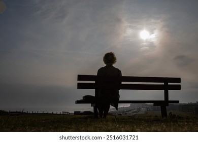 A lonely mature woman sits on a bench, on a hillside overlooking a coastal town. She is viewed from the rear, in near silhouette in late afternoon sun. The length of empty bench emphasises loneliness.