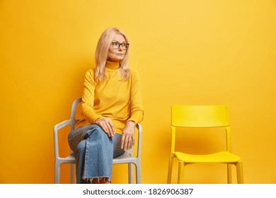 Lonely Middle Aged Woman Sits Near Empty Chair Needs Communication Looks Thoughtfully Away Dressed In Casual Turtleneck And Jeans Isolated On Yellow Background. Getting Older And Depression.