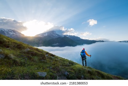 Lonely Mountaineer Descending From Kazbek 5054m Mountain With Backpack, Helmets High Mountaineering Boots And Trekking Poles Standing And Enjoying Sunset After Successful Ascending, Caucasus, Georgia.