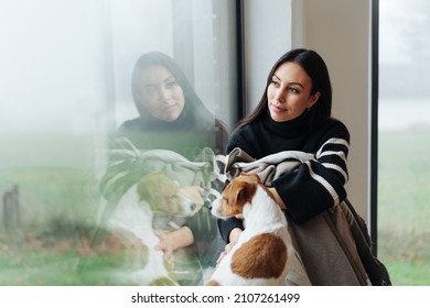 Lonely And Nostalgic Young Woman Looking Away Through The Window While Sitting Next To Her Cute Dog At Home In A Foggy Autumn Day