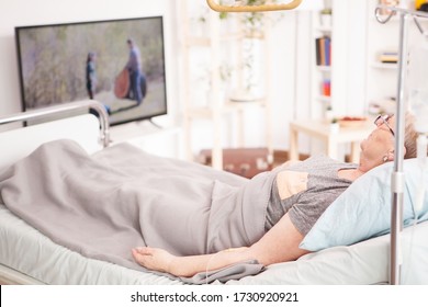 Lonely old woman in nursing home lying in bed watching tv.