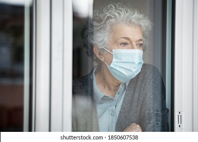 Lonely old woman wearing surgical mask and looking through the window during lockdown. Senior sad woman with face protective mask stay at home. Depressed lady at home during the covid-19 pandemic. - Powered by Shutterstock