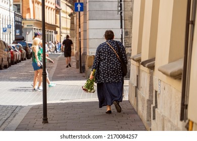 A Lonely Overweight Woman Walks With A Bouquet Of Flowers Along The Sidewalk Of An Old European City From The Shadows To The Light, Along Which A Couple Of A Woman And A Man Walk