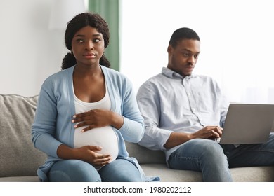 Lonely pregnant african american woman sitting by her ignoring husband using laptop, chatting with his friends. Sad expecting black lady looking for attention from her man, living room interior