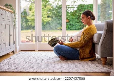 Lonely Pregnant Woman Sitting On Floor At Home Holding Army Husband's Cap
