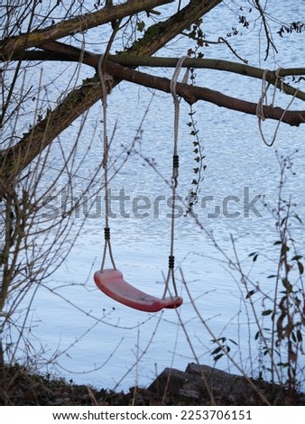 A lonely red child swing hangs on a branch dangerously close to the edge of a river