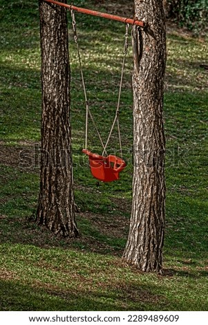 Lonely red children's swing in the garden with big conifer trees against green grass background. Dry brush effect