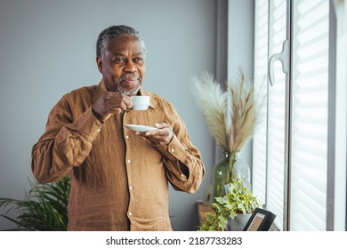 A lonely retired old African man with a white cup of coffee in his hands looks out the window and stands behind the glass during isolation. Concept coronavirus pandemic. - Powered by Shutterstock