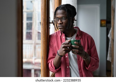 Lonely sad African man looking out window standing indoors suffering due to separation from girlfriend or lack of friends. Unhappy young black guy in casual clothes holding mug with tea or coffee - Shutterstock ID 2232388935