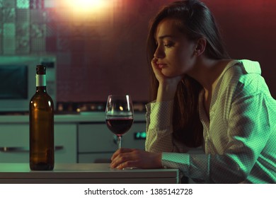 Lonely, Sad, Beautiful Young Woman With Red Wine Is Drinking Alone In The Kitchen At Home In Evening. Female Alcoholism And Alcohol Addiction. 