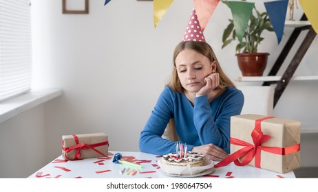 A Lonely Sad Girl Business Woman Sits At A Table With A Cake And Gifts With Her Head In A Cap On Her Hand. A Bored Young Lady Is At Home At A Party Alone. Remote Birthday Concept. Place For Text