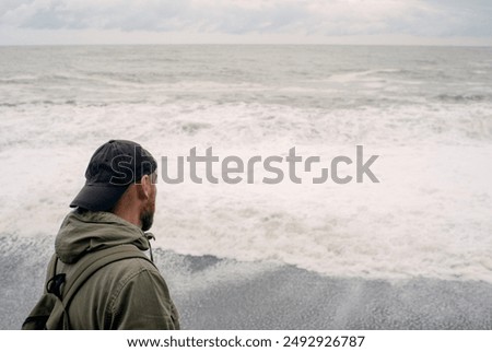 A lonely sad man in a cap and with a backpack looks into the distance at the stormy sea on a cloudy day in autumn