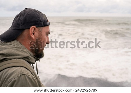 A lonely sad man in a cap and with a backpack looks into the distance at the stormy sea on a cloudy day in autumn